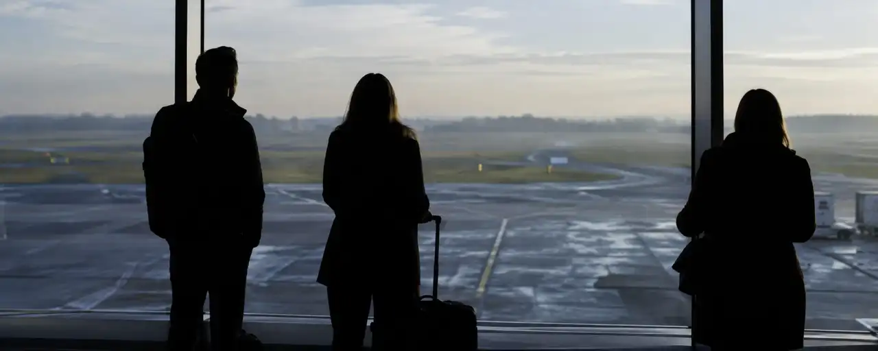 Three travellers in silhouette at an airport window, looking out at the runway